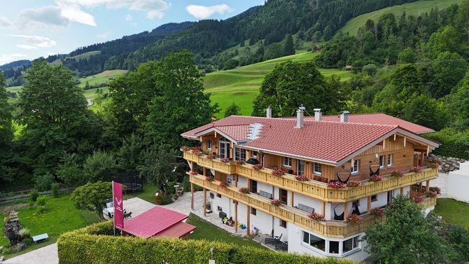 Ein großes Holzhaus ist von Bäumen, Wiesen und Bergen umgeben.