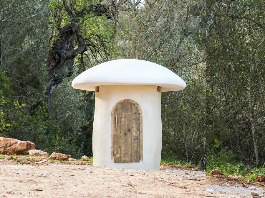 Un edificio blanco con forma de hongo con una puerta de madera en medio de un bosque.