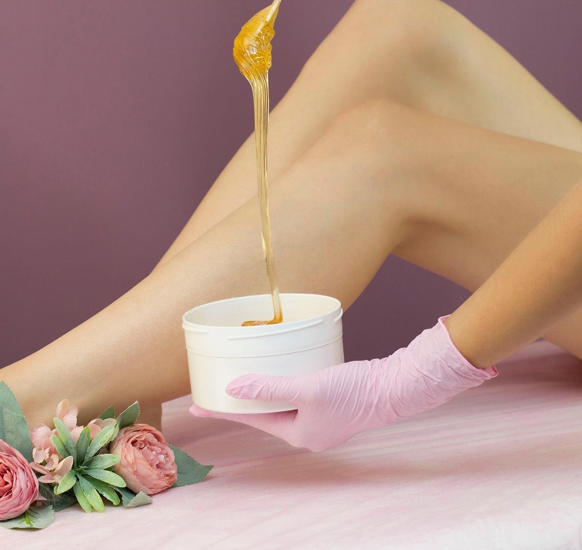 Pierna de una persona siendo depilada con cera. Una mano enguantada sostiene un recipiente de cera. La cera ámbar gotea sobre la pierna.