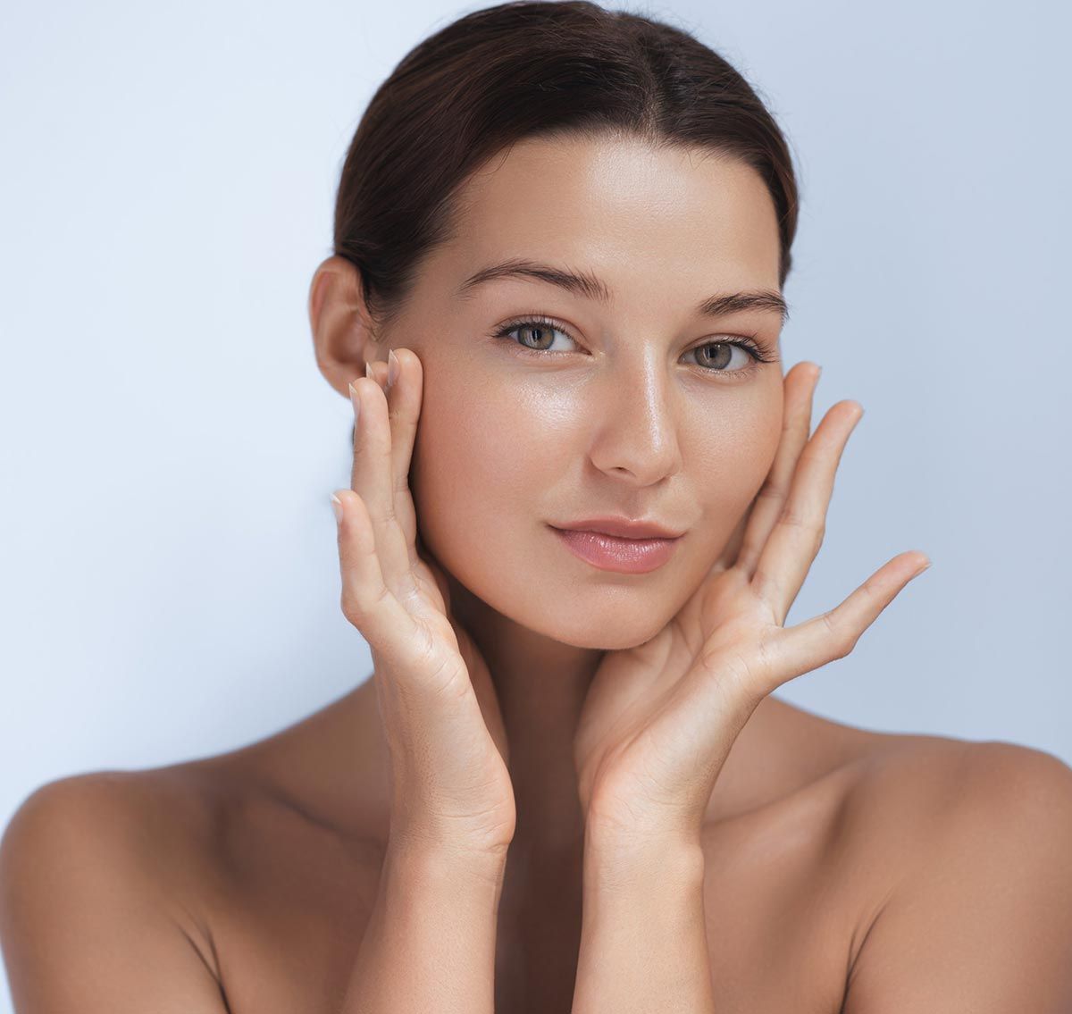 Mujer con las manos en la cara, mirando a la cámara. La piel se ve tersa, el fondo es azul claro.