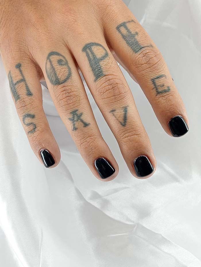 Mano con uñas negras y tatuajes en los dedos que forman la frase