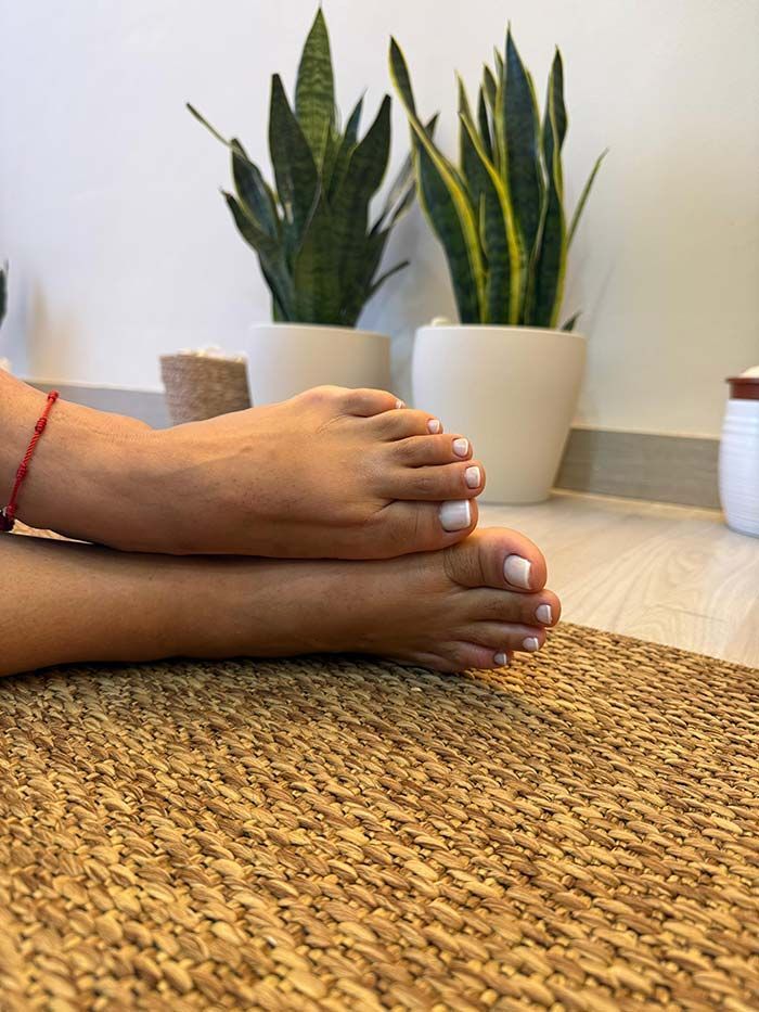 Pies descalzos con uñas pintadas de blanco, cruzados sobre una esterilla de yoga de corcho, contra una pared blanca con plantas en macetas.