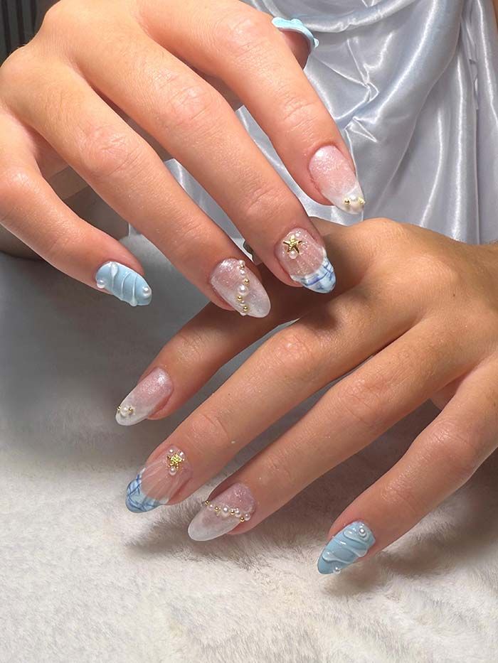 Manos con diseños de uñas en azul pastel y transparente, decoradas con gemas y perlas.