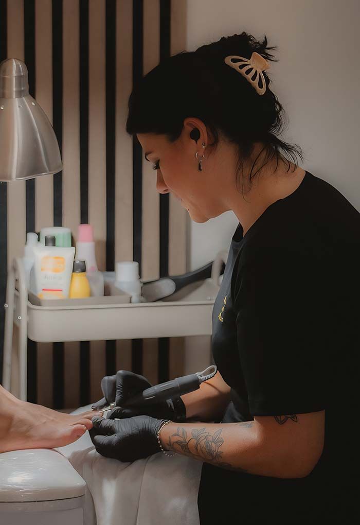 Mujer realizando una pedicura con un taladro en un salón de belleza. Lleva guantes y camisa negros.