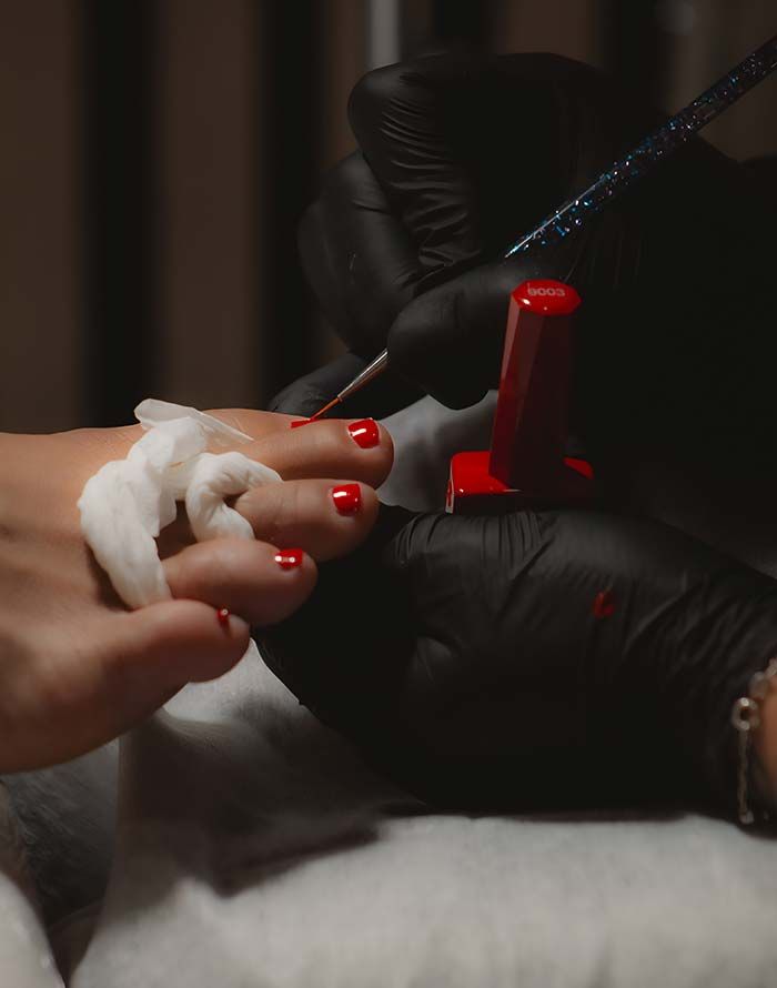 Pedicurista aplicando esmalte de uñas rojo en los dedos de los pies de una clienta, con separadores de algodón colocados.