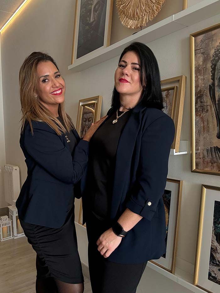 Dos mujeres con blazers azul marino están de pie una al lado de la otra, sonriendo, frente a una pared con cuadros enmarcados.