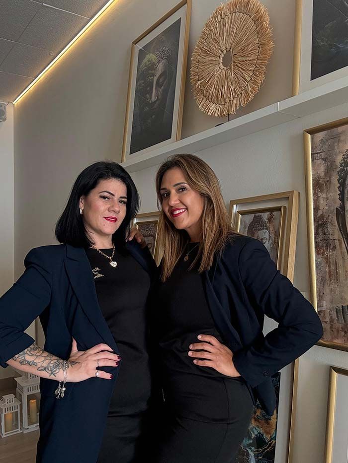 Dos mujeres con blazers oscuros posan frente a una obra de arte. Una tiene tatuajes y ambas sonríen con las manos en las caderas.