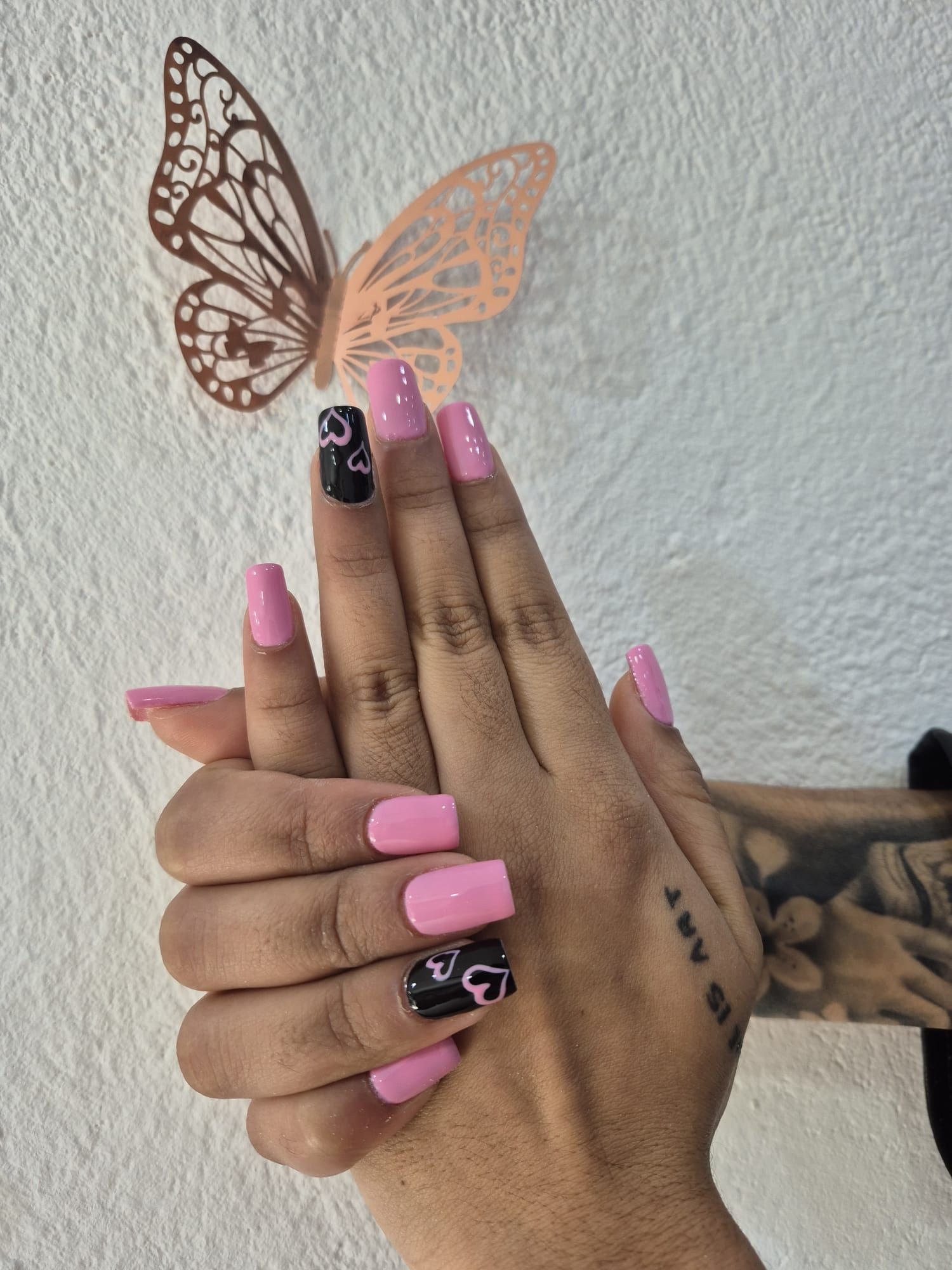 Una mano de mujer con uñas rosas y negras y una mariposa en la pared detrás de ella.