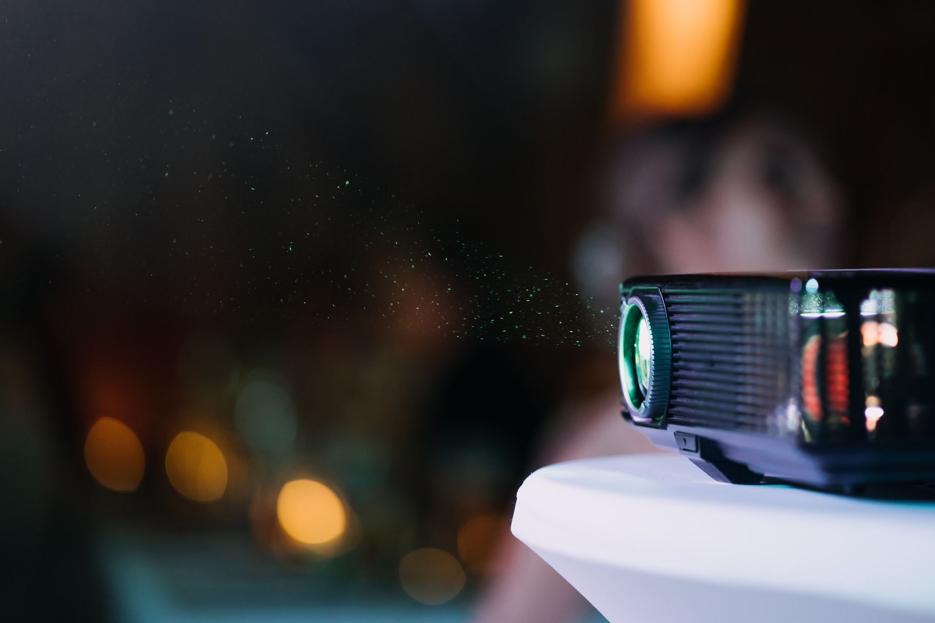 Proyector negro sobre una mesa blanca proyectando una luz con partículas en una habitación oscura con luces de fondo borrosas.