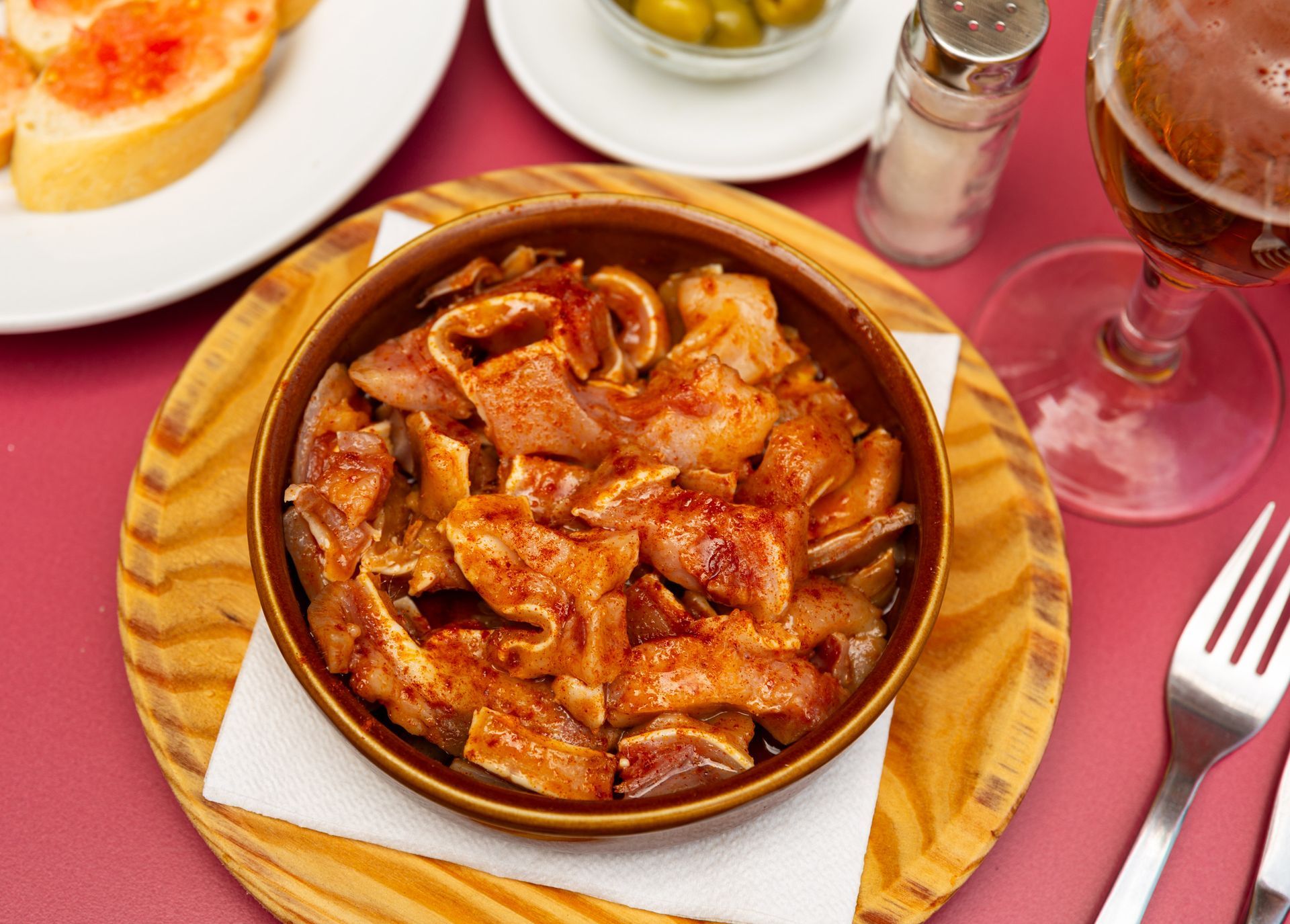Primer plano de un plato de tapas de oreja de cerdo frita sazonada con pimentón, en un plato de madera, con pan, aceitunas y cerveza.