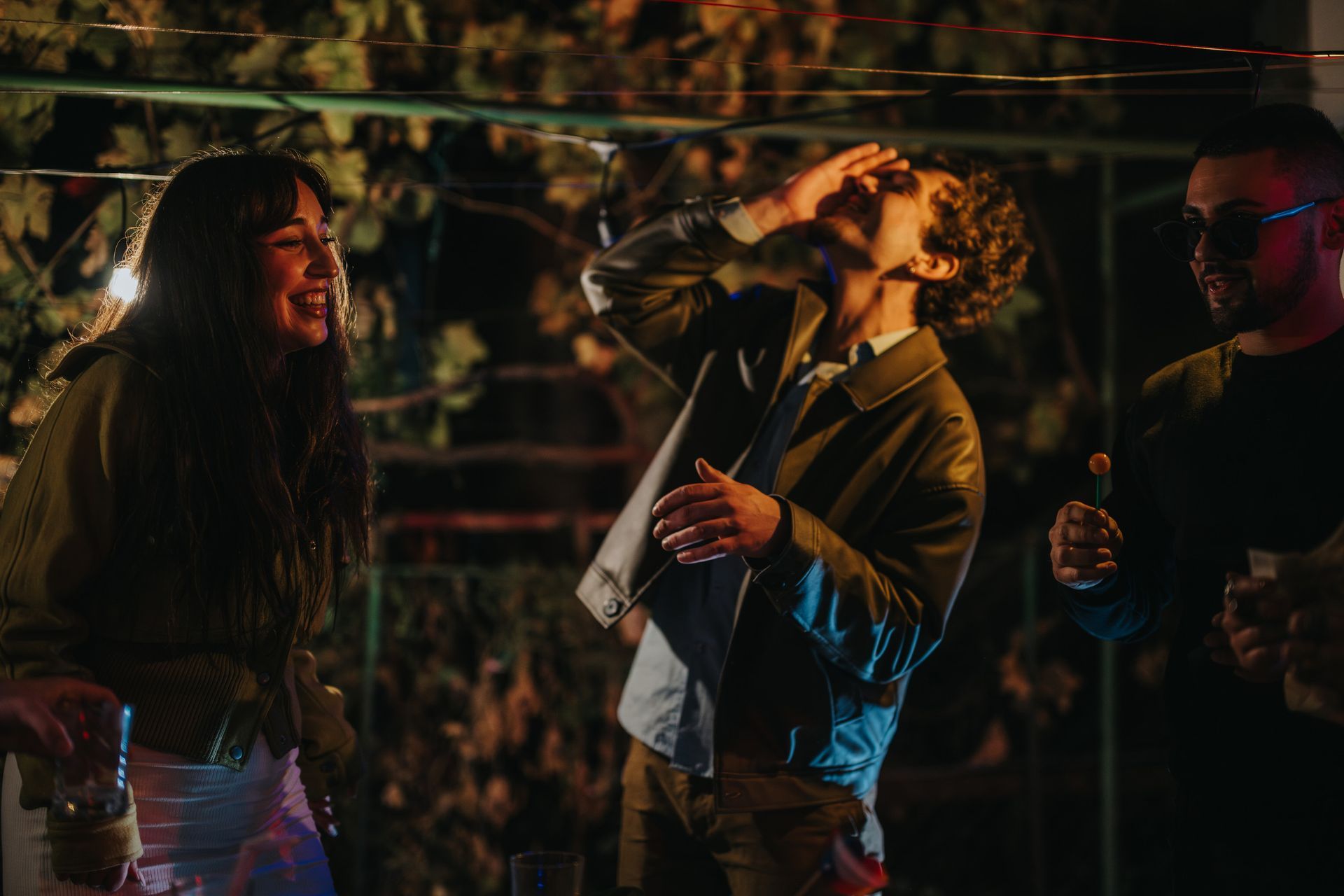 Tres personas en una fiesta al aire libre: una bebiendo, las demás riendo. Escenario nocturno con guirnaldas de luces y enredaderas.