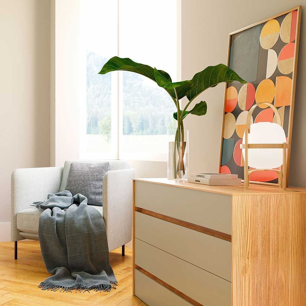 Una habitación bien iluminada con un sillón gris claro, una cómoda de madera, obras de arte de gran tamaño y plantas.