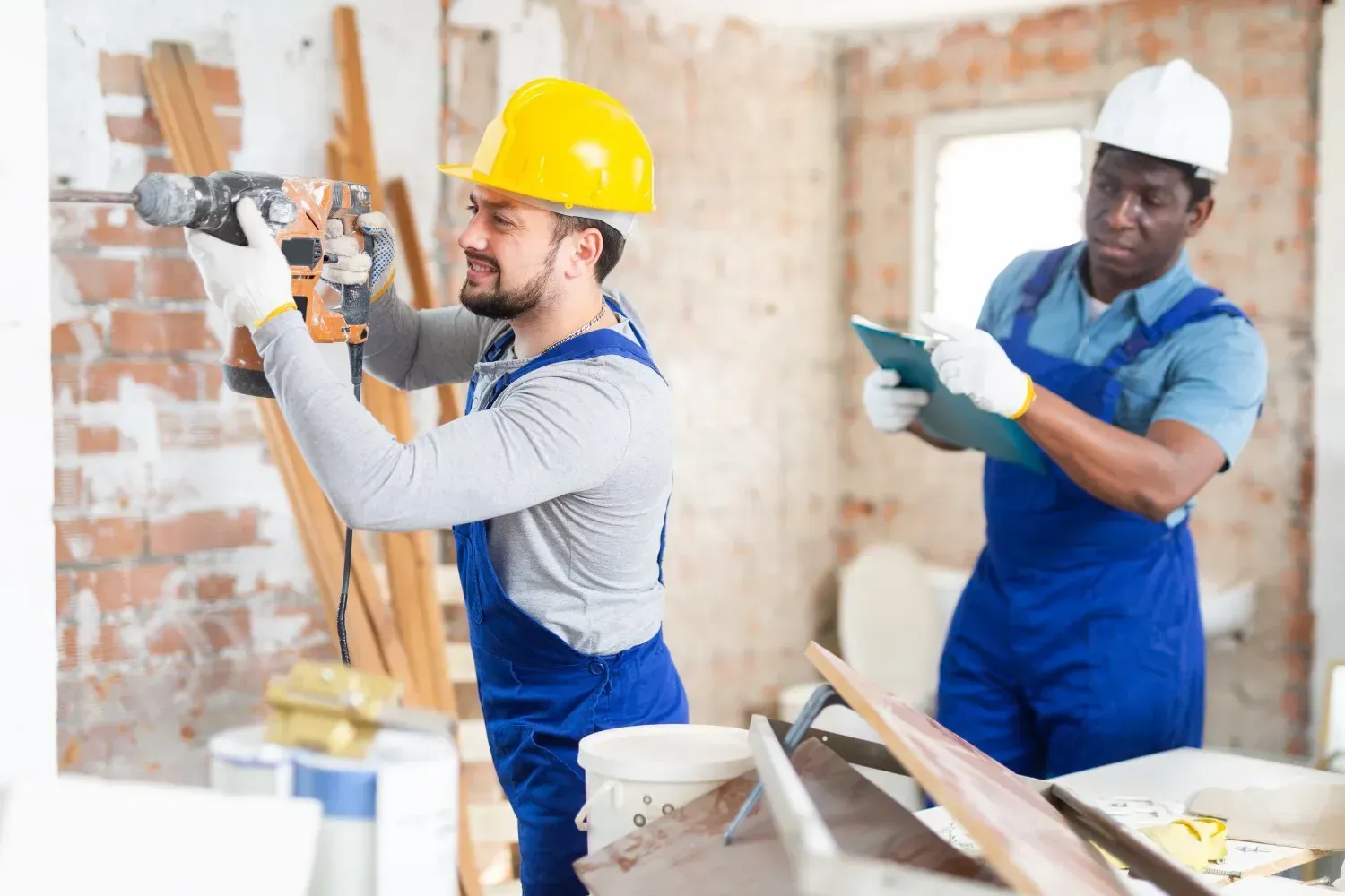Dos obreros de la construcción: uno perforando una pared, el otro sujetando un portapapeles. Interior de una obra.