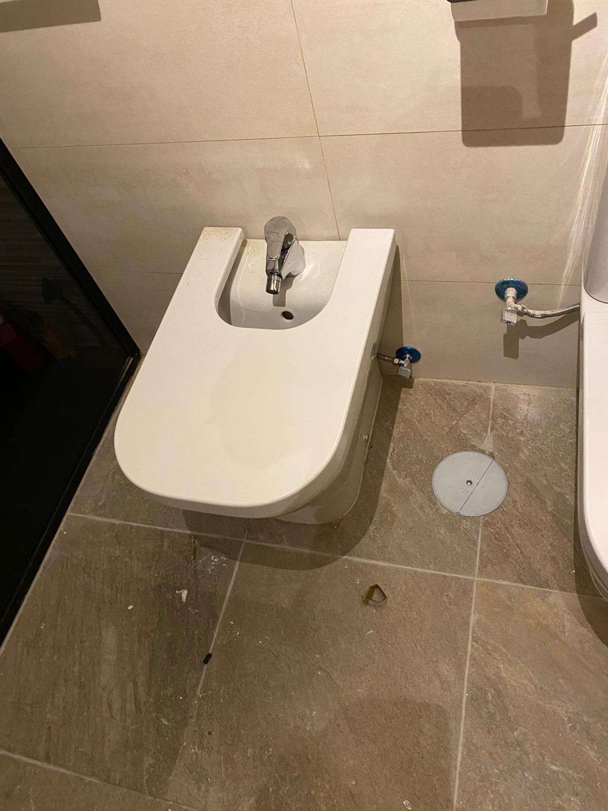 Bidé blanco en un baño con suelo de baldosas beige y grifo de pared.