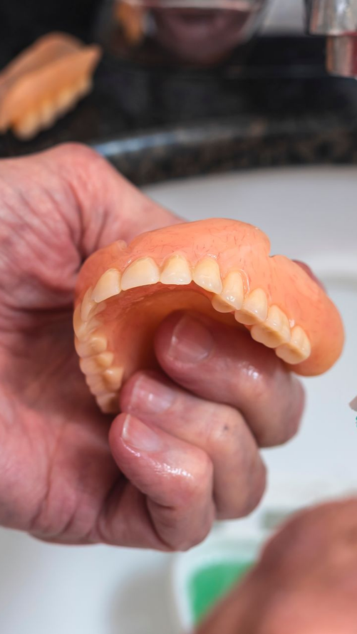 Personne tenant un dentier en main près d'un évier ; couleurs pêche et blanc.