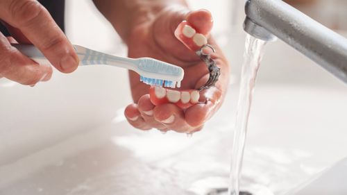 Une personne brosse ses prothèses dentaires sous l'eau courante dans un évier blanc.