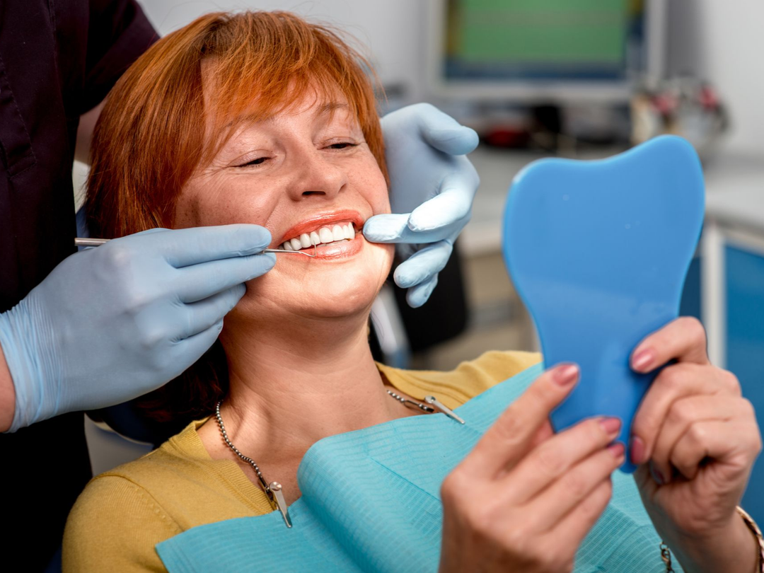 Un dentiste examine les dents d'une femme avec un miroir. La femme sourit, assise sur un fauteuil, vêtue d'un t-shirt jaune.