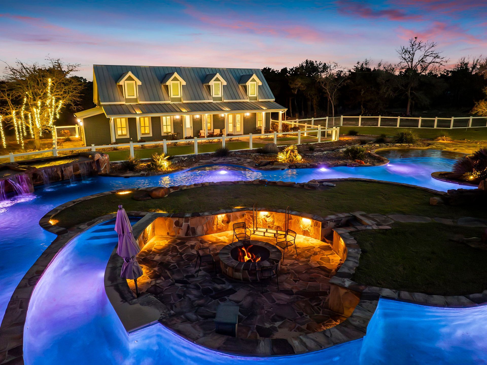 Luxury home with pool, fire pit, and outdoor kitchen, illuminated at dusk.