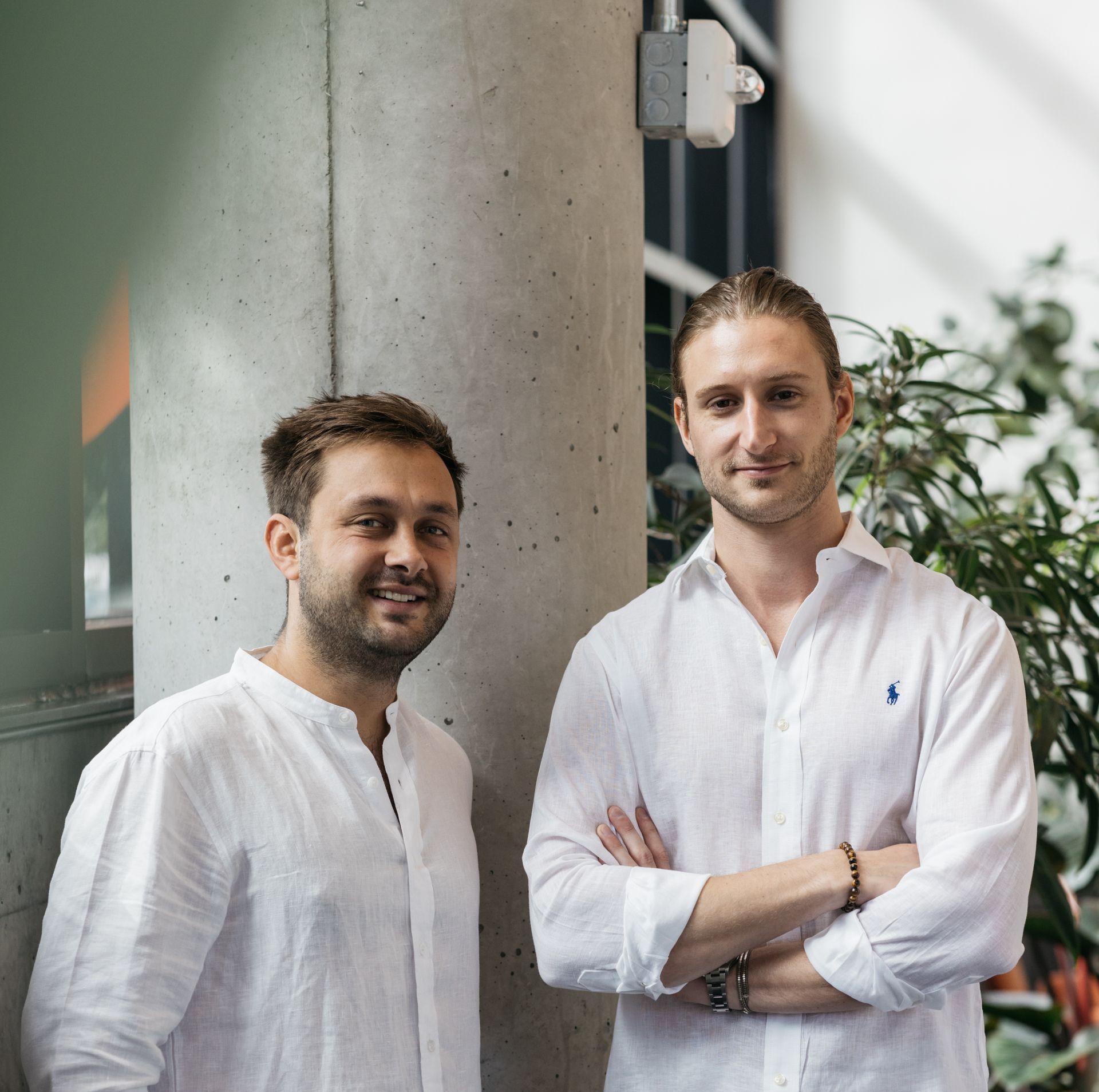 Two men smiling, dressed in white, standing next to a pillar in a bright, modern space.