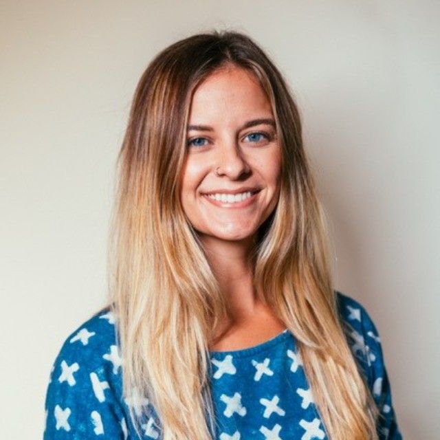 Woman with blonde hair smiling, wearing blue patterned top, neutral background.