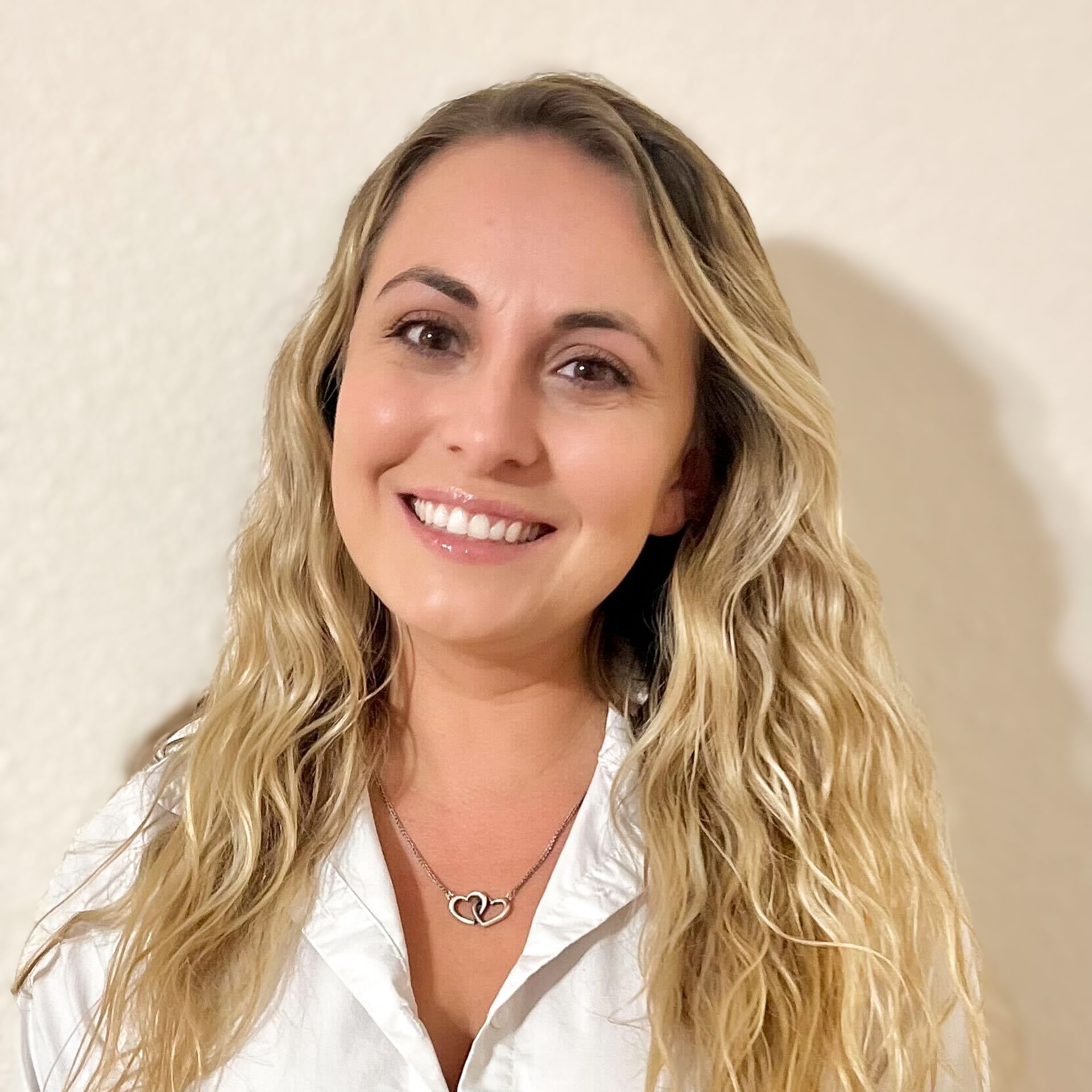 Woman with long wavy blonde hair smiles, wearing a white shirt and silver heart necklace.