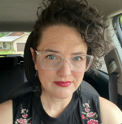 Woman with curly hair and glasses in a car, wearing a floral top and lipstick.