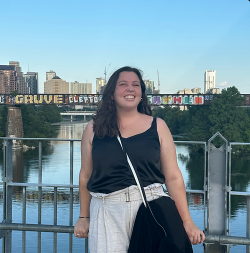 Woman smiles on a bridge in Austin, TX. She wears black top, white pants, and has a crossbody bag; city skyline background.