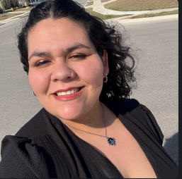 Woman with dark curly hair smiles at the camera, wearing a black top, silver necklace, and standing on a sunny street.