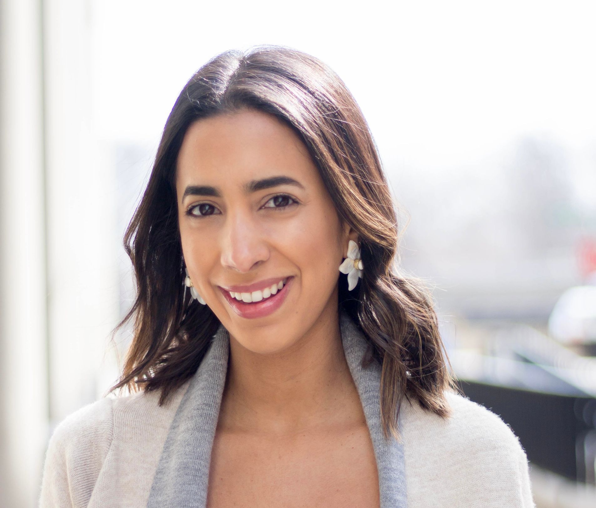 Woman with brown hair smiles, wearing a light cardigan and earrings. Outdoor setting, bright.