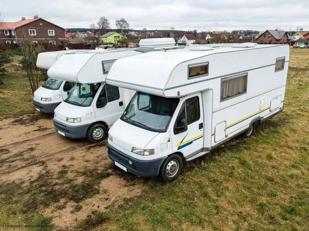 Drei weiße Wohnmobile parken nebeneinander auf einer Wiese, im Hintergrund sind Häuser zu sehen.