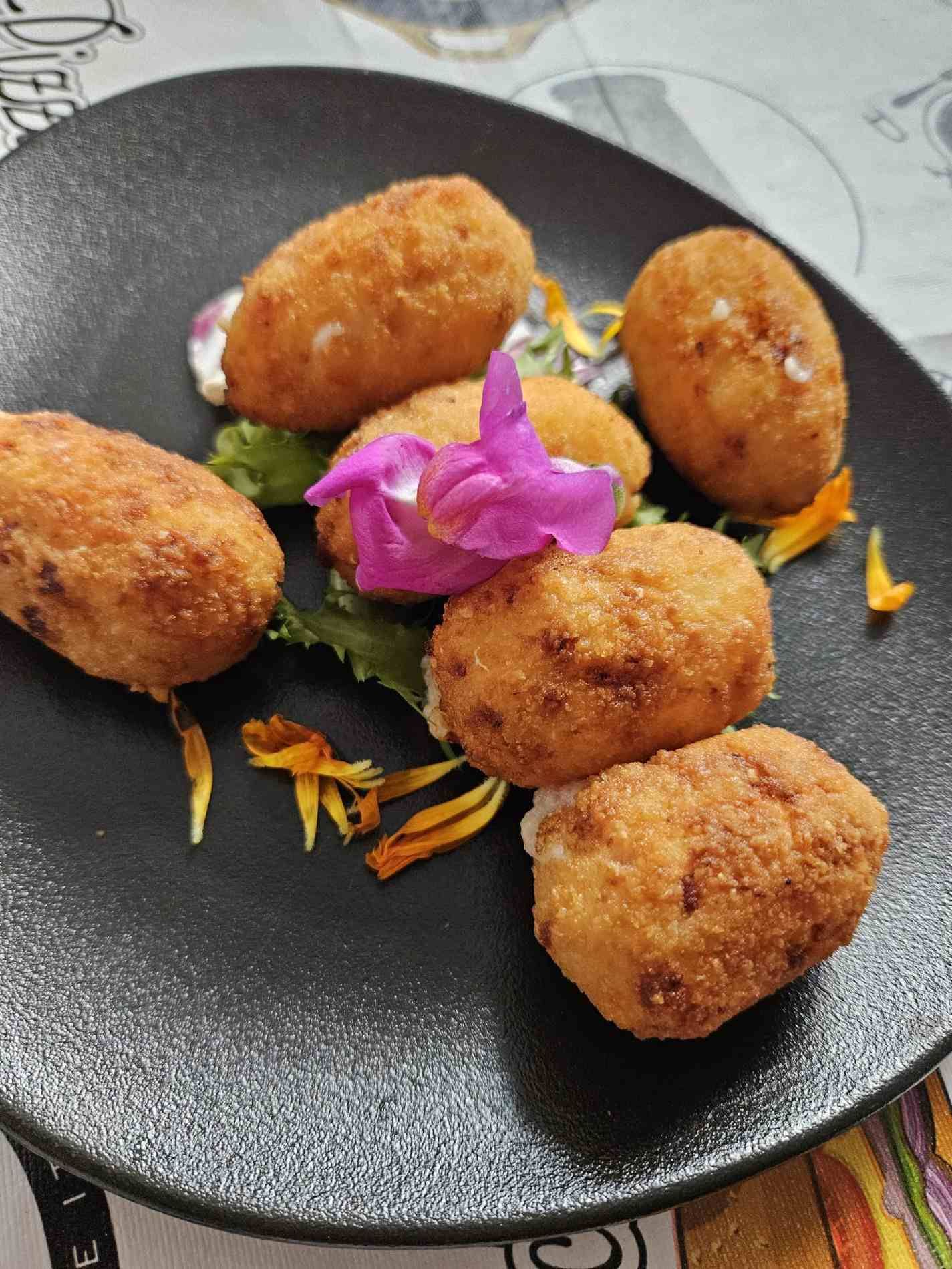 Croquetas fritas, doradas, dispuestas en un plato negro con una flor decorativa y guarnición.