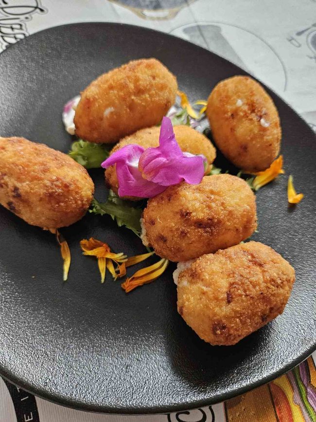 Croquetas fritas, doradas, dispuestas en un plato negro con una flor decorativa y guarnición.