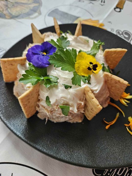 Primer plano de una ensalada compuesta en un plato negro, cubierta con flores comestibles, perejil y galletas.