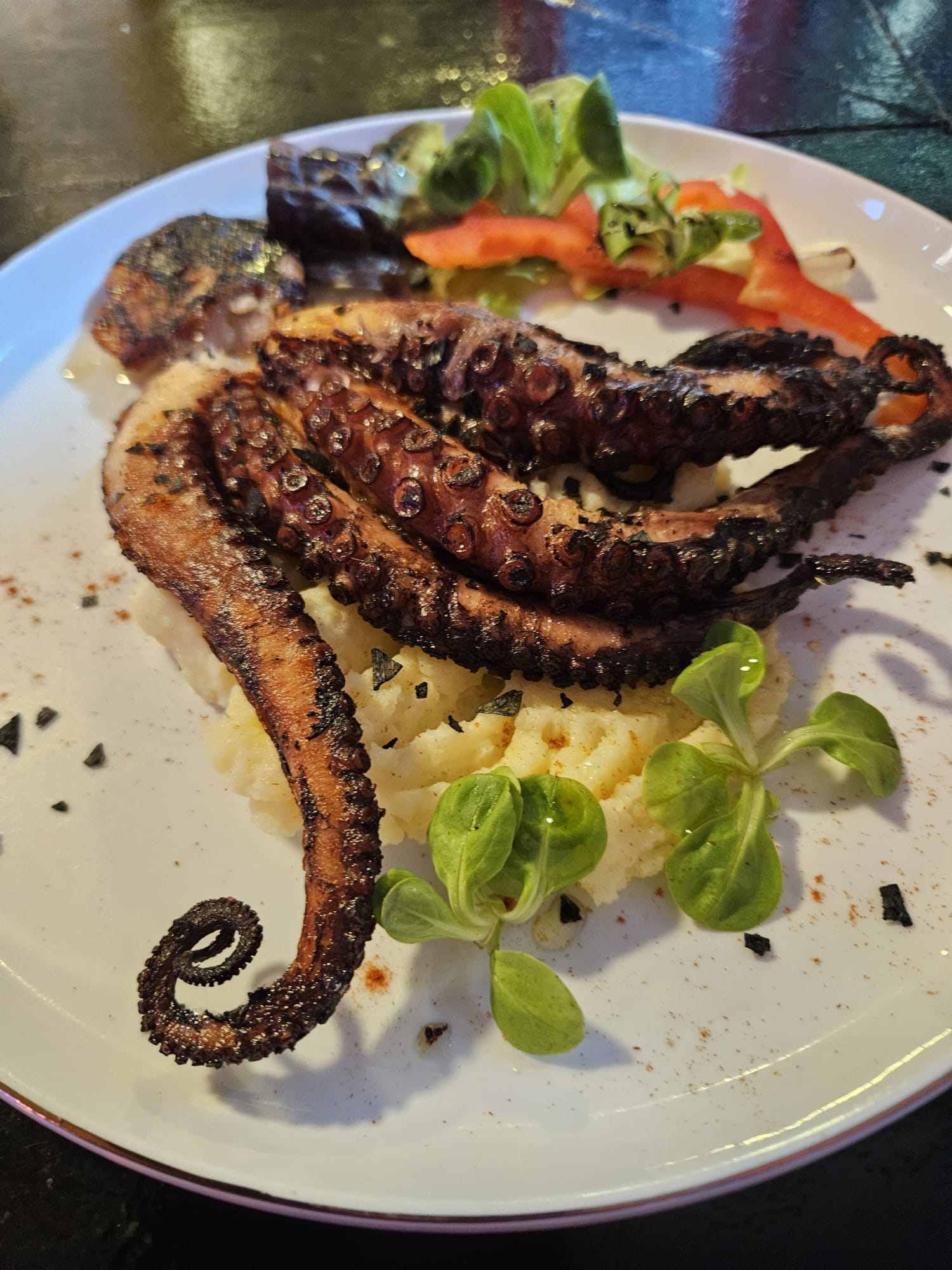 Pulpo a la parrilla sobre puré de patatas, adornado con verduras y hortalizas en un plato blanco.