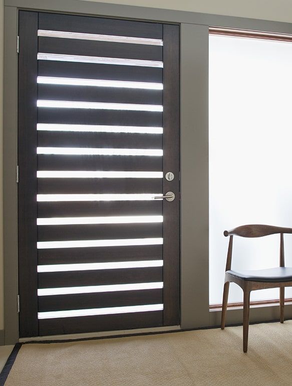 Porte d'entrée en bois foncé avec panneaux vitrés horizontaux. Une chaise est posée à droite, près d'une fenêtre.