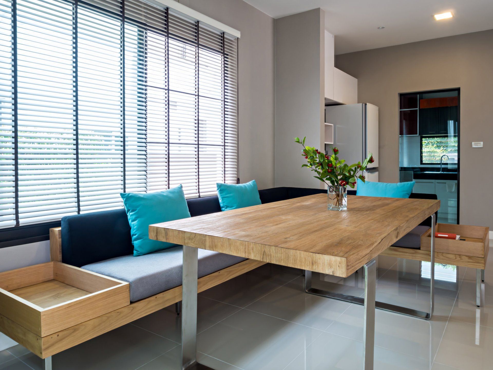 Coin repas avec une table en bois, un canapé avec des coussins bleu sarcelle et une grande fenêtre avec stores.