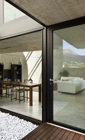 Les portes vitrées s'ouvrent sur l'intérieur avec une salle à manger et un salon, des murs en béton et une vue extérieure sur les montagnes.