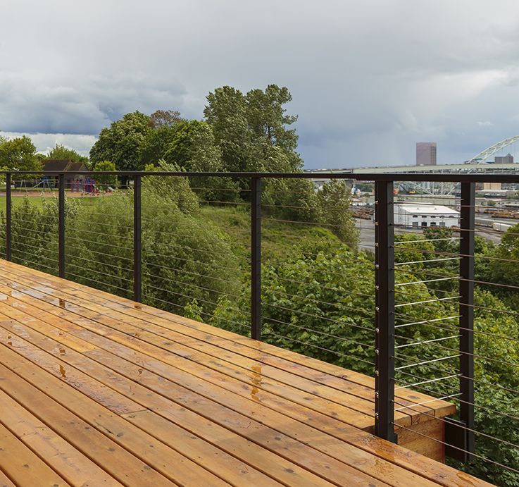 Terrasse en bois avec garde-corps en câble donnant sur un paysage urbain et des arbres verts sous un ciel nuageux.