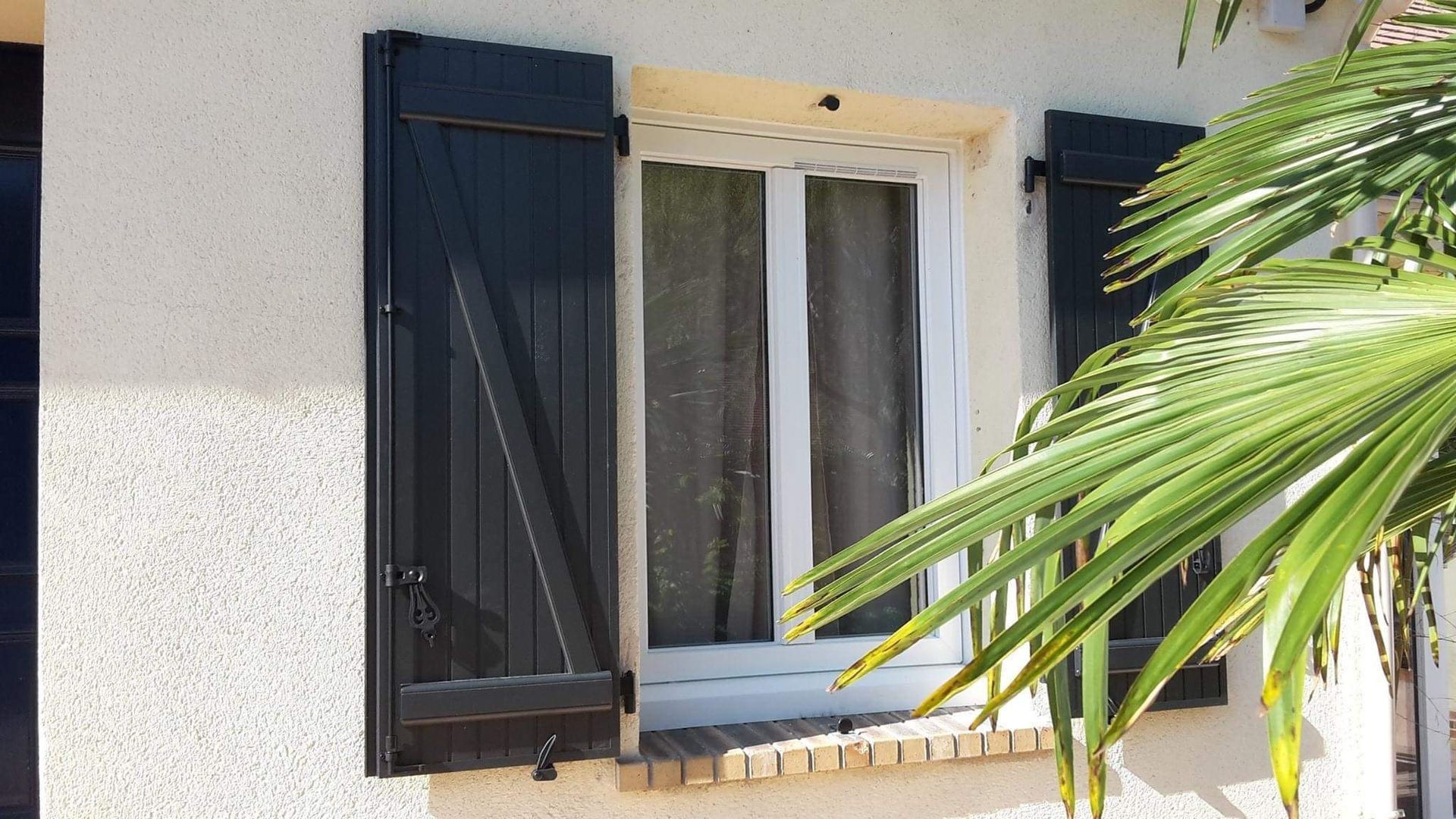 Fenêtre en bois avec volets ouverts sur un mur en stuc beige, baigné de soleil.