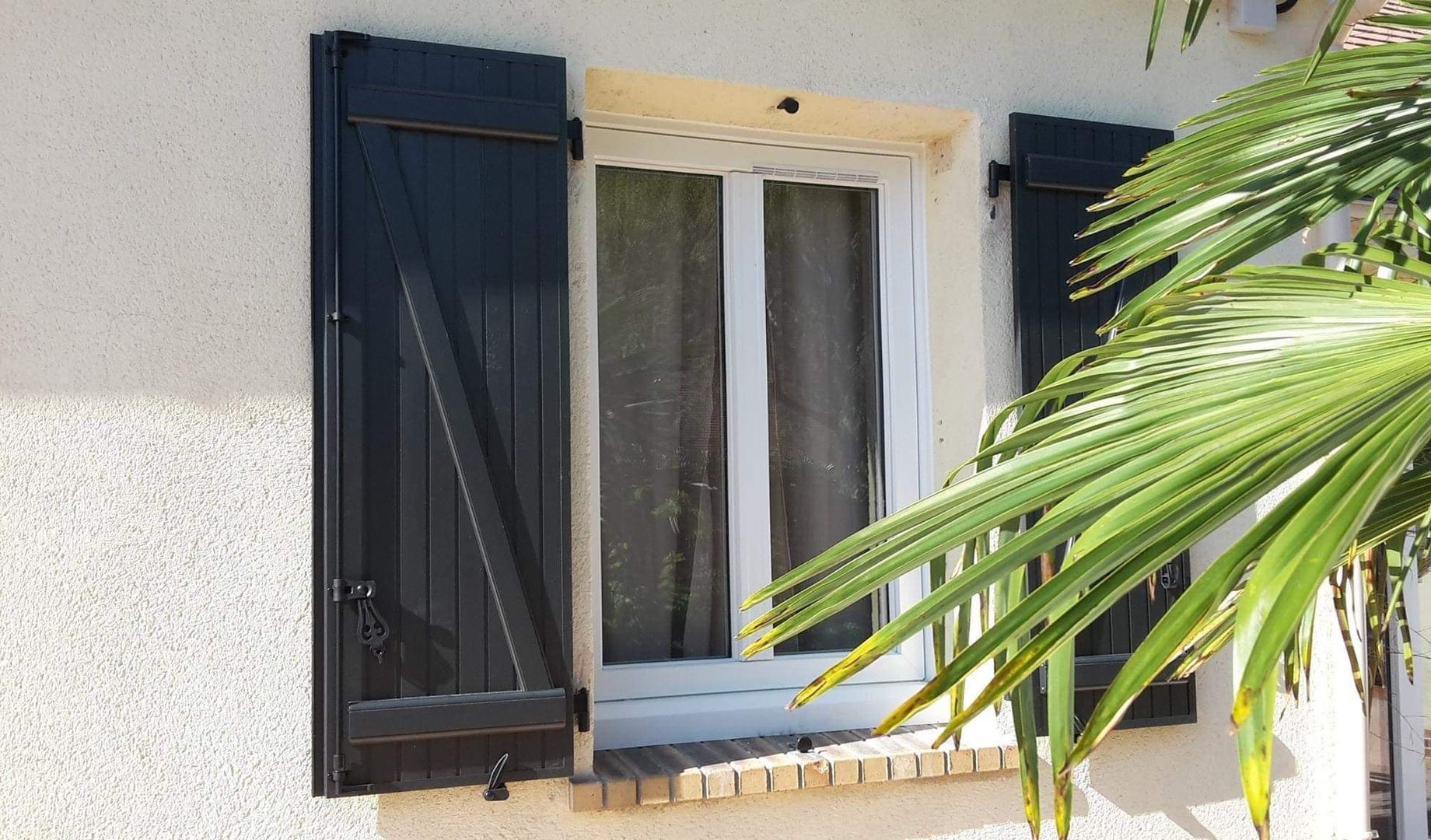 Fenêtre en bois avec volets ouverts sur un mur en stuc beige, baigné de soleil.