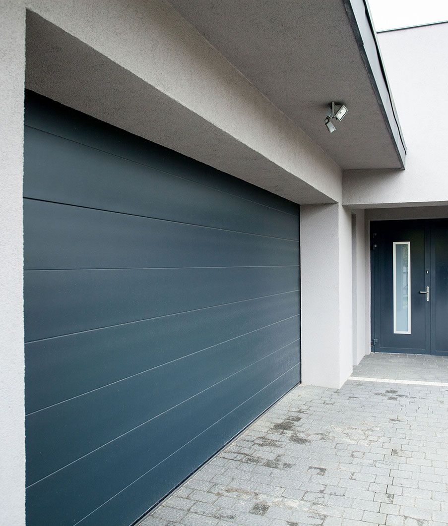 Porte de garage grise à côté d'une porte foncée, toutes deux sous un surplomb blanc.
