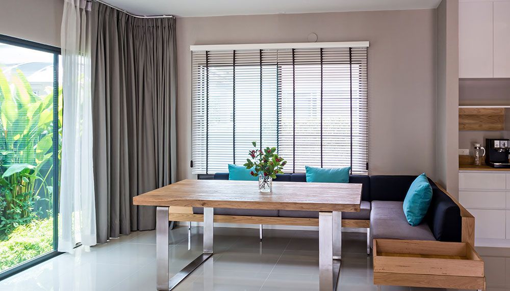 Coin repas : table en bois, banc en L avec coussins bleus, grande fenêtre avec verdure, rideaux gris.