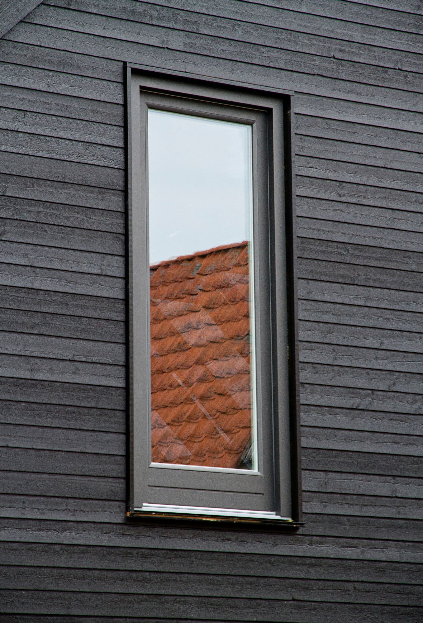 Longue fenêtre étroite dans un cadre gris reflétant un toit de tuiles rouges, insérée dans un mur horizontal en bois gris foncé.