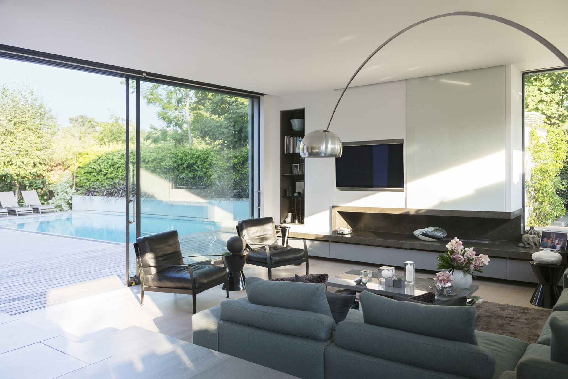 Salon moderne avec vue sur la piscine grâce à de grandes baies vitrées coulissantes. Canapé et fauteuils gris, décoration moderne et épurée.