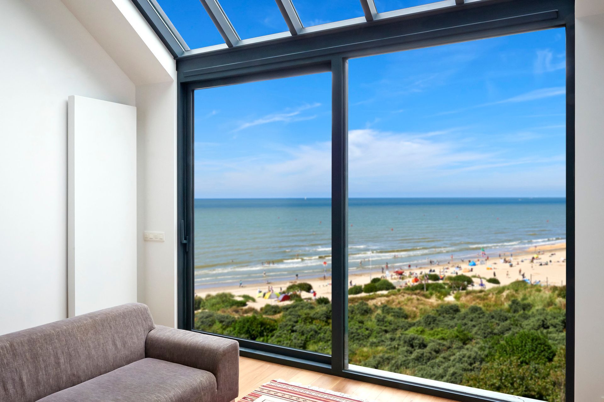 Chambre intérieure avec canapé et grande fenêtre donnant sur la plage et l'océan bleu. Ciel ensoleillé.