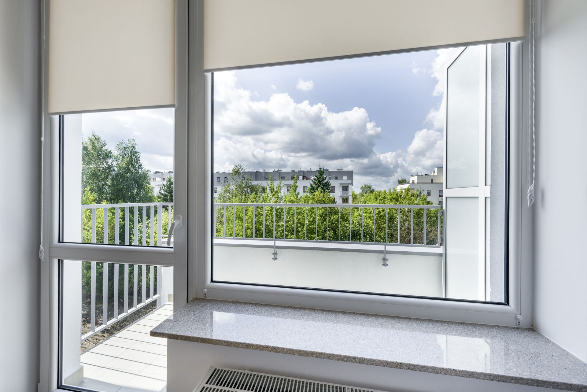 Fenêtre avec vue sur balcon et arbres, ciel bleu avec nuages.