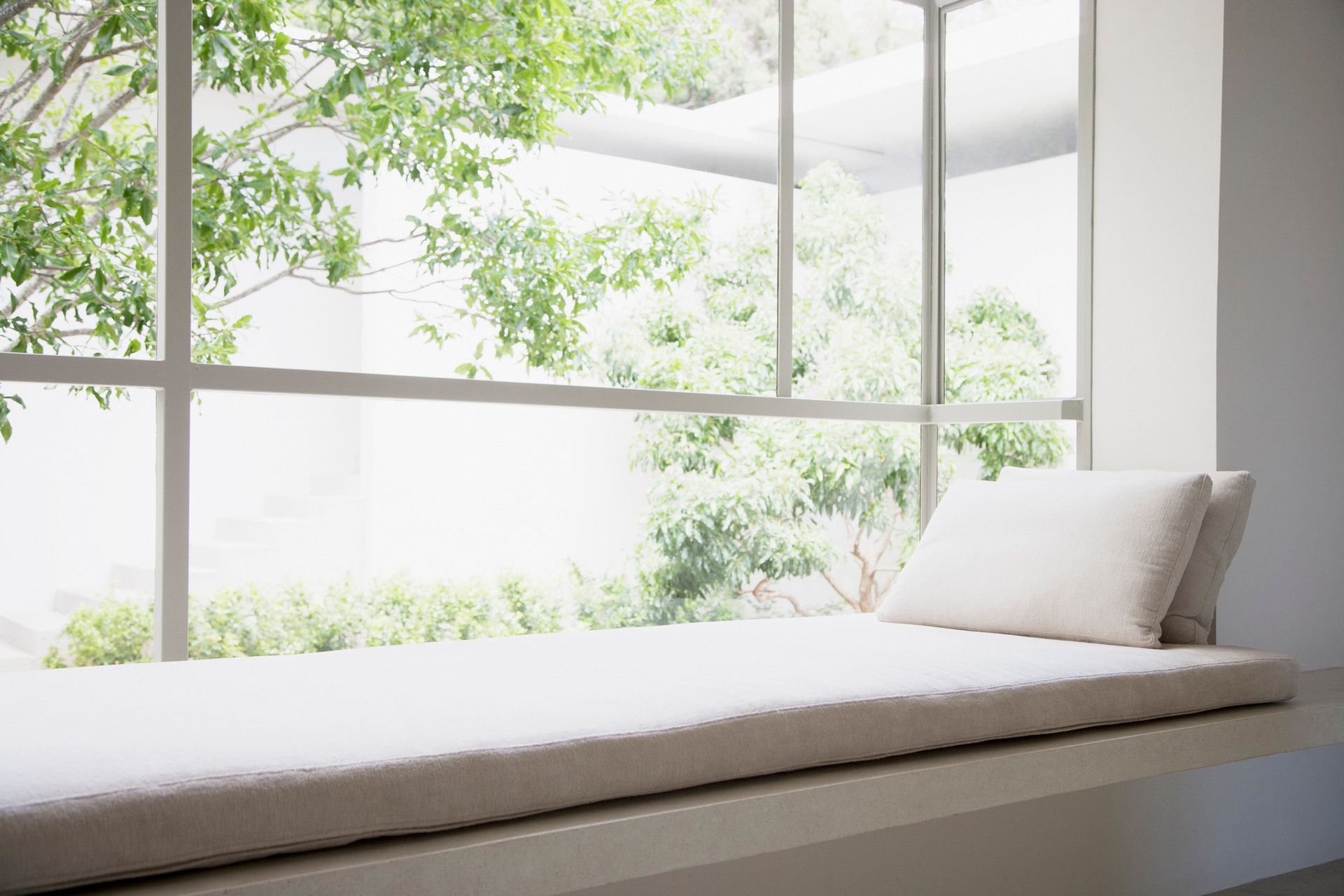 Banquette de fenêtre blanche avec coussin, donnant sur un feuillage vert luxuriant.