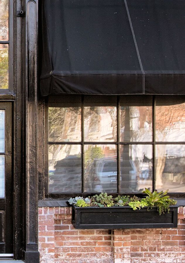 Auvent noir au-dessus d'une fenêtre avec un cadre noir et une base en brique ; une jardinière avec des plantes se trouve en dessous.