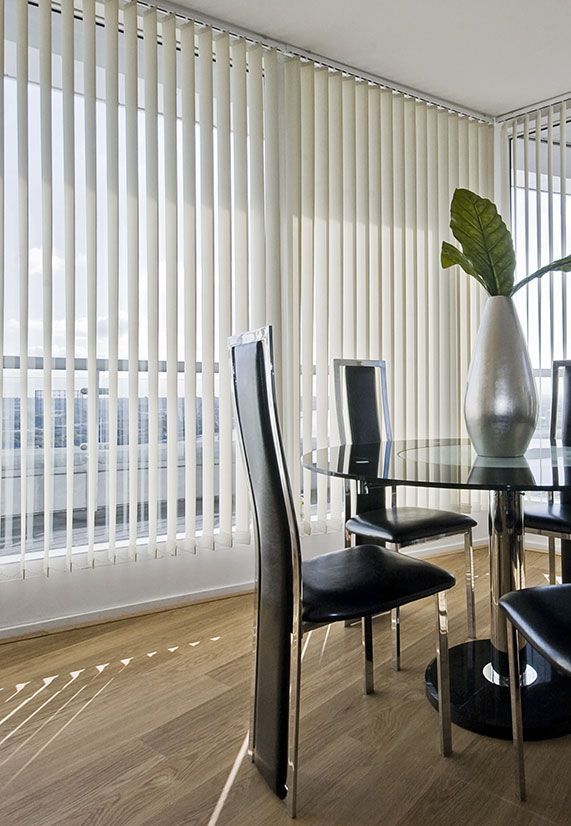 Salle à manger avec stores verticaux, table en verre, chaises noires et un vase argenté.
