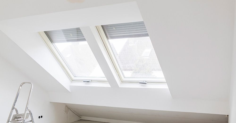 Deux fenêtres de toit blanches dans un plafond blanc en angle, avec des stores gris fermés. Un escabeau se trouve dans le coin.